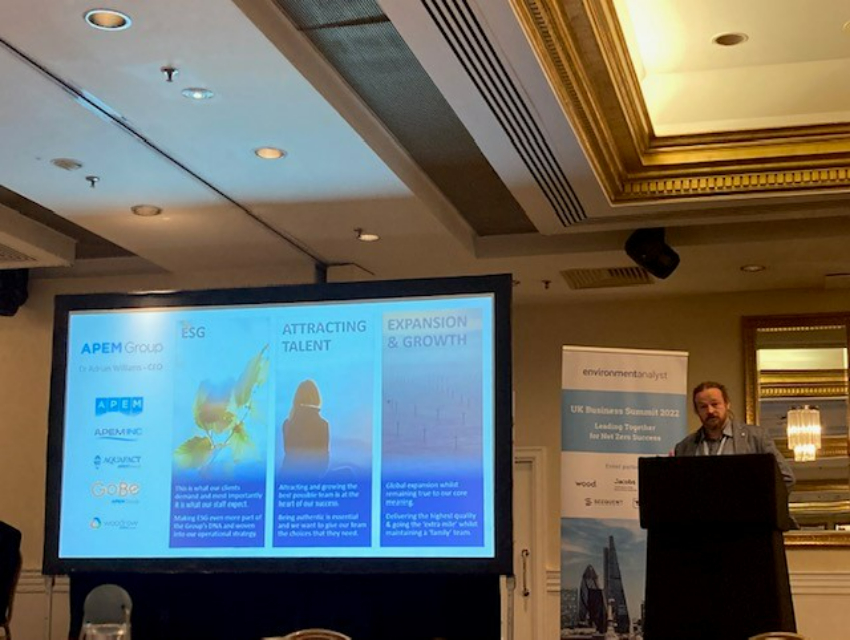 A man standing behind a lectern in a conference room. Behind him is a banner and to his right is a screen showing a presentation by the APEM Group. The subtitles are ESG, Attracting Talent and Expansion & Growth